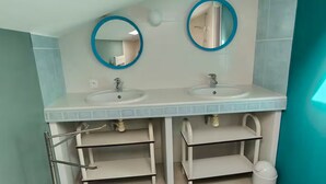 Une salle de bain avec deux lavabos, chacun étant surmonté d’un miroir. On trouve des étagères sous les lavabos et une poubelle sur la droite.