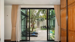 A bedroom with a bed, a view of a pool, and a glass door leading to an outdoor area with a lounge chair and a table. - Wings 25 Laguna (Phuket)