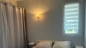 A bedroom with a bed, a nightstand, a wall-mounted light, a window with blinds, and a curtain. - House near the beach (Saint-Georges-d'Oléron)