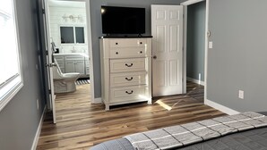 Un dormitorio con cama, cómoda, televisor y baño visible a través de una puerta abierta.