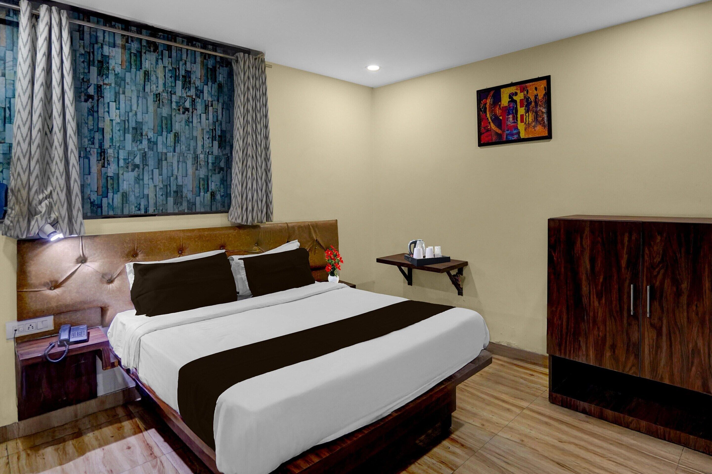 Una habitación de hotel con cama, mesita de noche, una pequeña mesa con hervidor, armario y un cuadro en la pared.