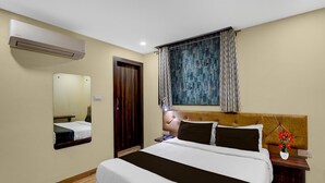 A hotel room with a bed, a bedside table, a nightstand, a mirror, an air conditioner, a window with curtains, and a wooden door.