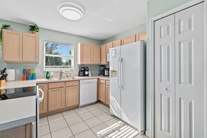 Une cuisine avec des meubles en bois, un réfrigérateur blanc, un lave-vaisselle et une fenêtre avec des stores.