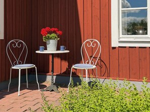 Zwei weiße Metallstühle mit gepolsterten Sitzen, ein kleiner runder Tisch mit einem Blumentopf und zwei Tassen, sowie ein Fenster mit Blick auf Grünflächen.