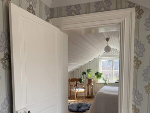Ein Zimmer mit hoher Decke, ein Holzstuhl, ein Blumentopf und ein Fenster mit Blick auf Bäume.