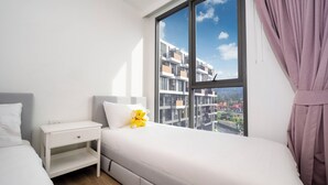Quarto de hotel moderno com uma janela grande, uma cama, uma mesinha de cabeceira e um urso de pelúcia.