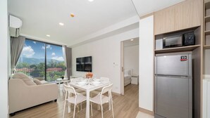 Uma sala de estar moderna com sofá, mesa de jantar, cadeiras, cozinha com geladeira e uma grande janela com vista para vegetação e montanhas.