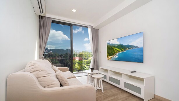 Uma sala de estar moderna com sofá, uma mesinha, TV na parede e uma varanda com vista para vegetação e montanhas.