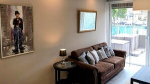 A modern living room with a brown leather sofa, a small side table, a framed picture, and a large window with a view of a pool and buildings.