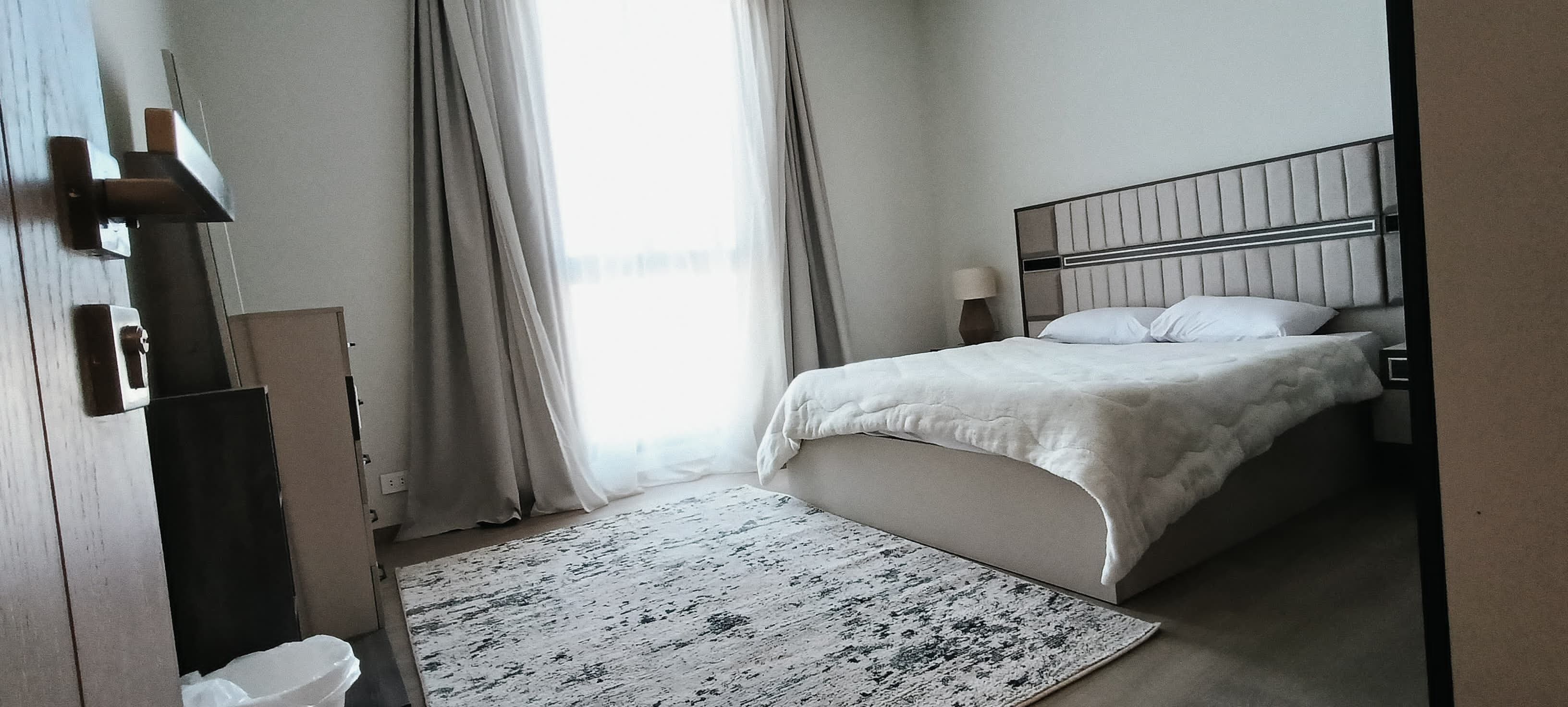 A bedroom with a bed, a nightstand, a lamp, a window with curtains, and a door.