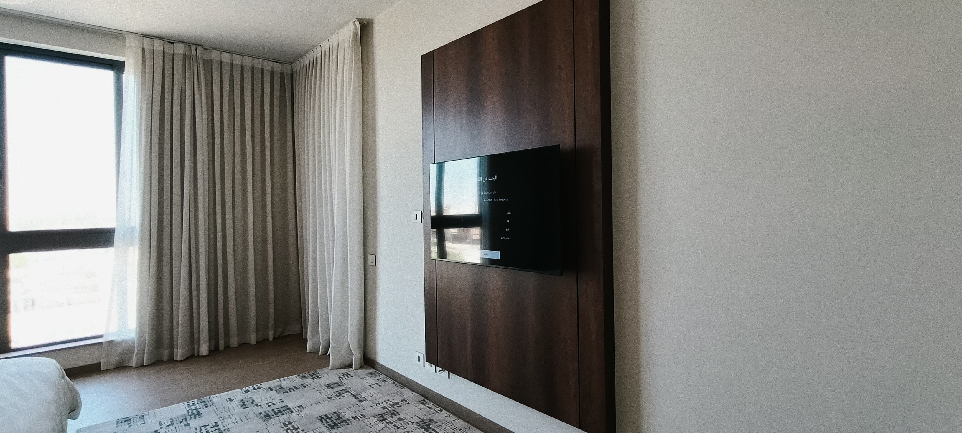 A modern hotel room with a large window, a flat-screen TV mounted on a wooden panel, and a patterned rug on the floor.