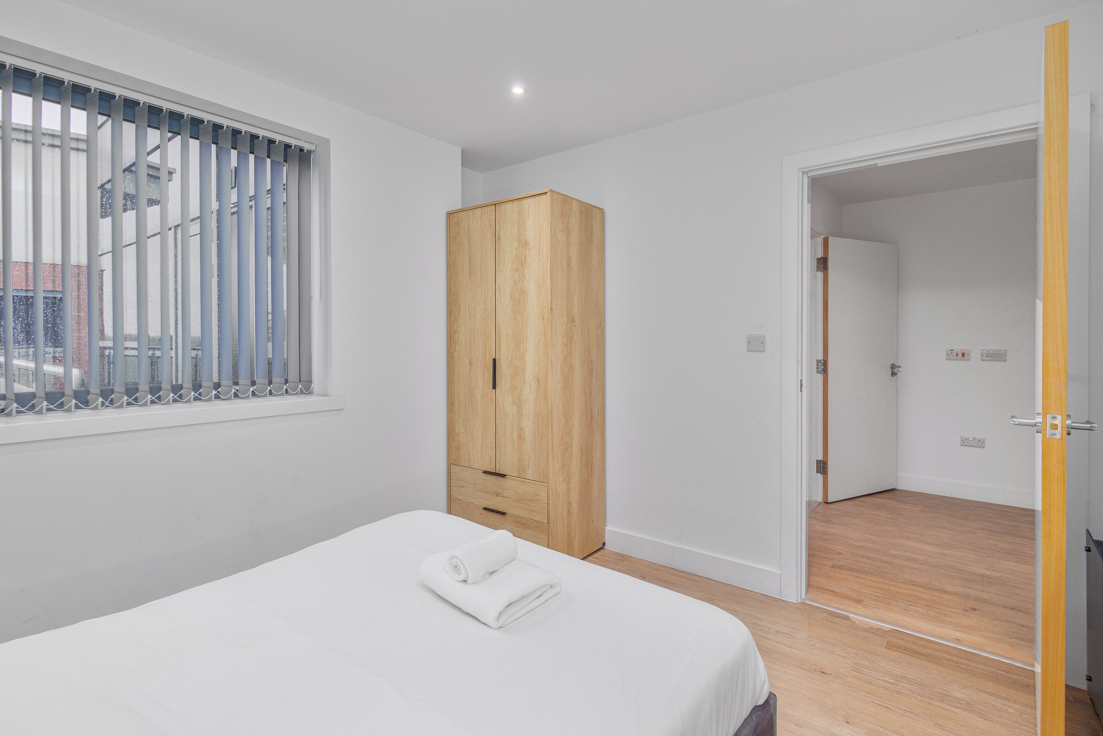 Een slaapkamer met een bed, een houten kledingkast, een raam met jaloezieën en een open deur naar een andere kamer.