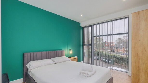 Ein modernes Schlafzimmer mit einem großen Bett, einem Nachttisch mit Lampe und Blick ins Freie durch ein großes Fenster.