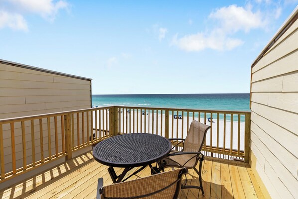 Eine Holzterrasse mit einem runden Tisch und zwei Stühlen mit Blick auf einen Strand, Menschen und einen klaren Himmel.