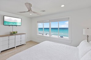 Ein Schlafzimmer mit einem Bett, einem Fernseher, einem Deckenventilator, einem Schminktisch und einem Fenster mit Blick auf den Strand.
