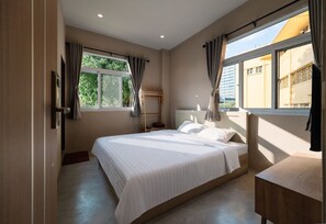 A bedroom with a bed, a bedside table, a window with curtains, and a view of buildings outside. - Kosin House (Bangkok)
