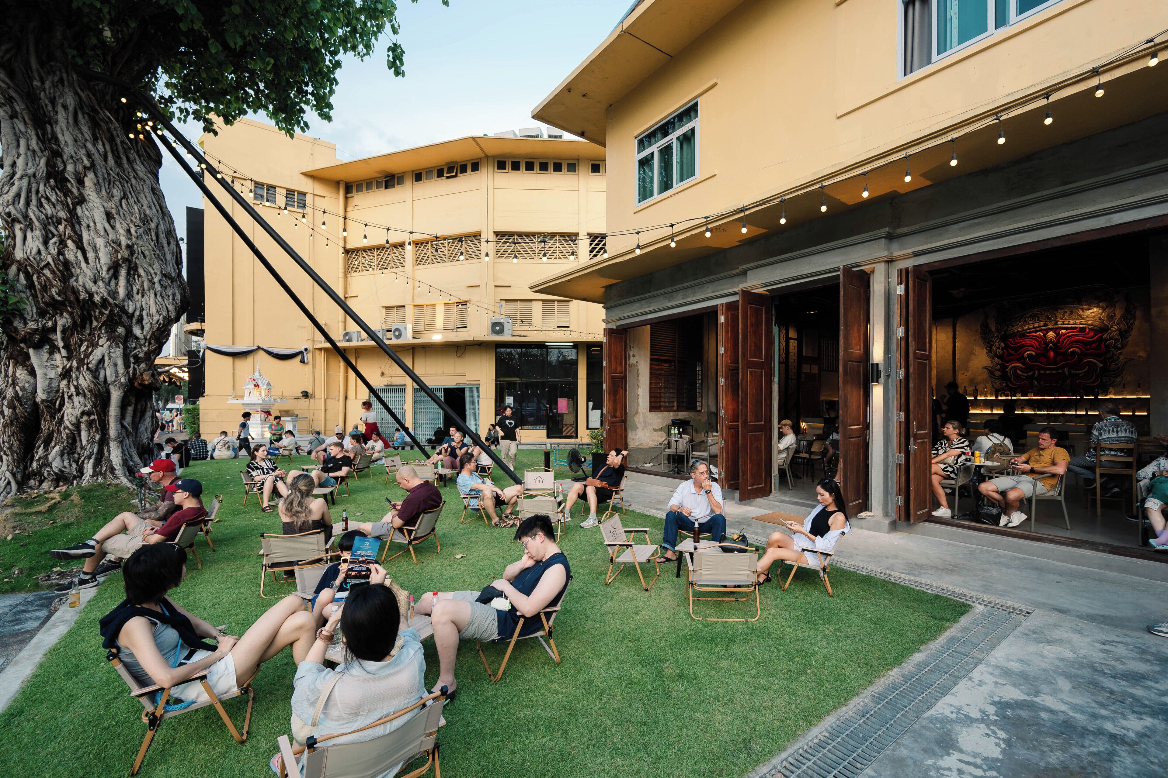 An outdoor seating area with people relaxing on chairs, a building with a balcony, and a restaurant entrance with a decorative facade.