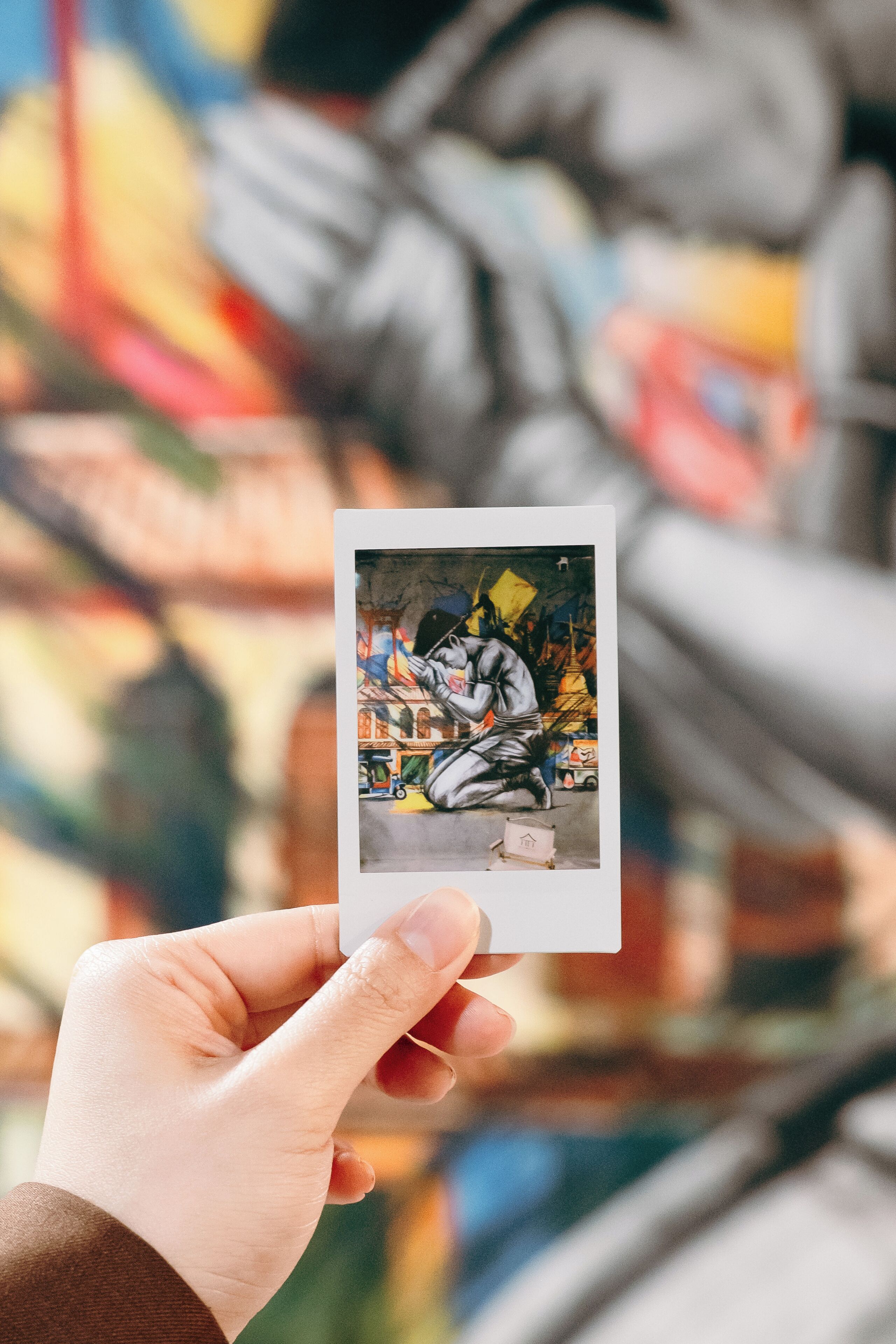 A hand holding a Polaroid photo of a person sitting on a bench with a colorful mural in the background.