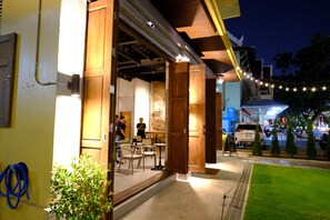 An open-air restaurant with wooden doors and tables, a visible outdoor area with greenery, and a street view with cars and a building in the background. - Kosin House (Bangkok)