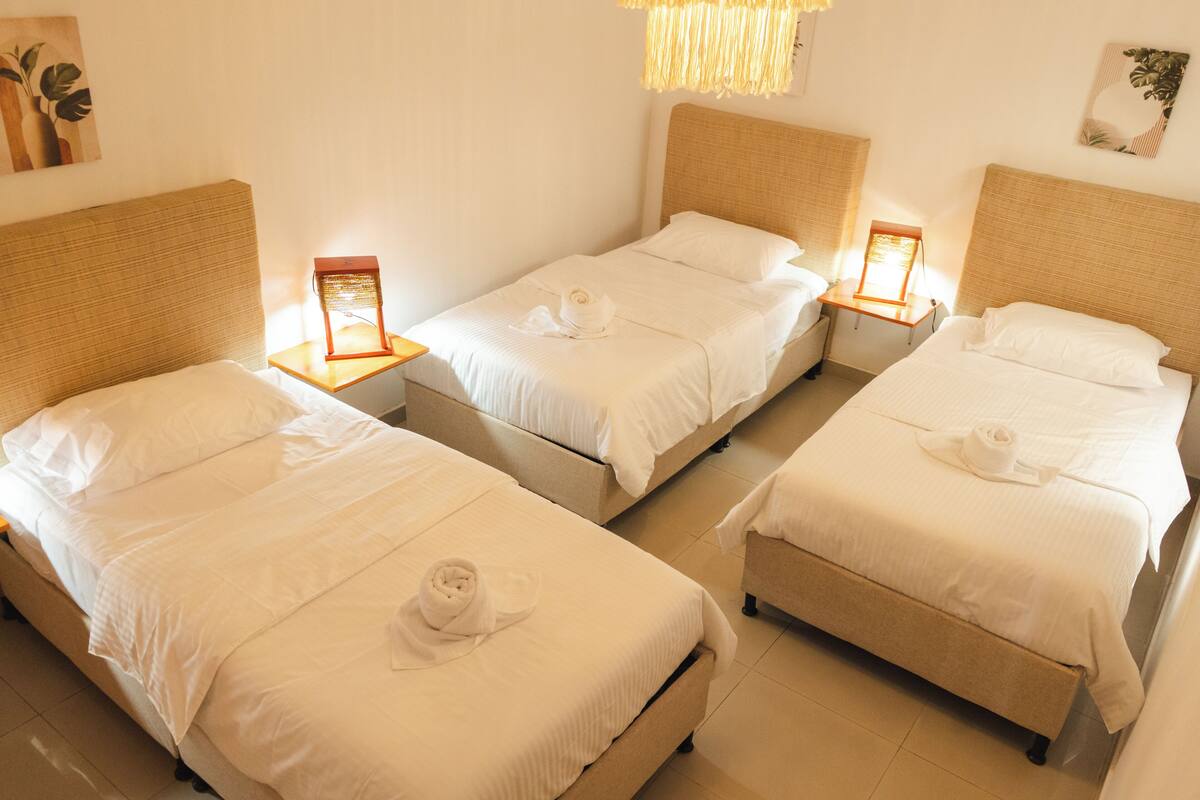 Habitación de hotel con tres camas, cada una con sábanas blancas y una toalla enrollada. Hay mesitas de noche con lámparas, y la habitación presenta un diseño moderno con una paleta de colores neutros.