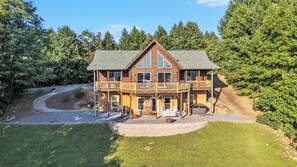 A large, two-story log cabin with a balcony, surrounded by a well-maintained lawn and dense forest. - Stunning Mountain Views & Sunsets, Hot Tub + Fire Pit,  4BR Blue Ridge Retreat (Nebo)