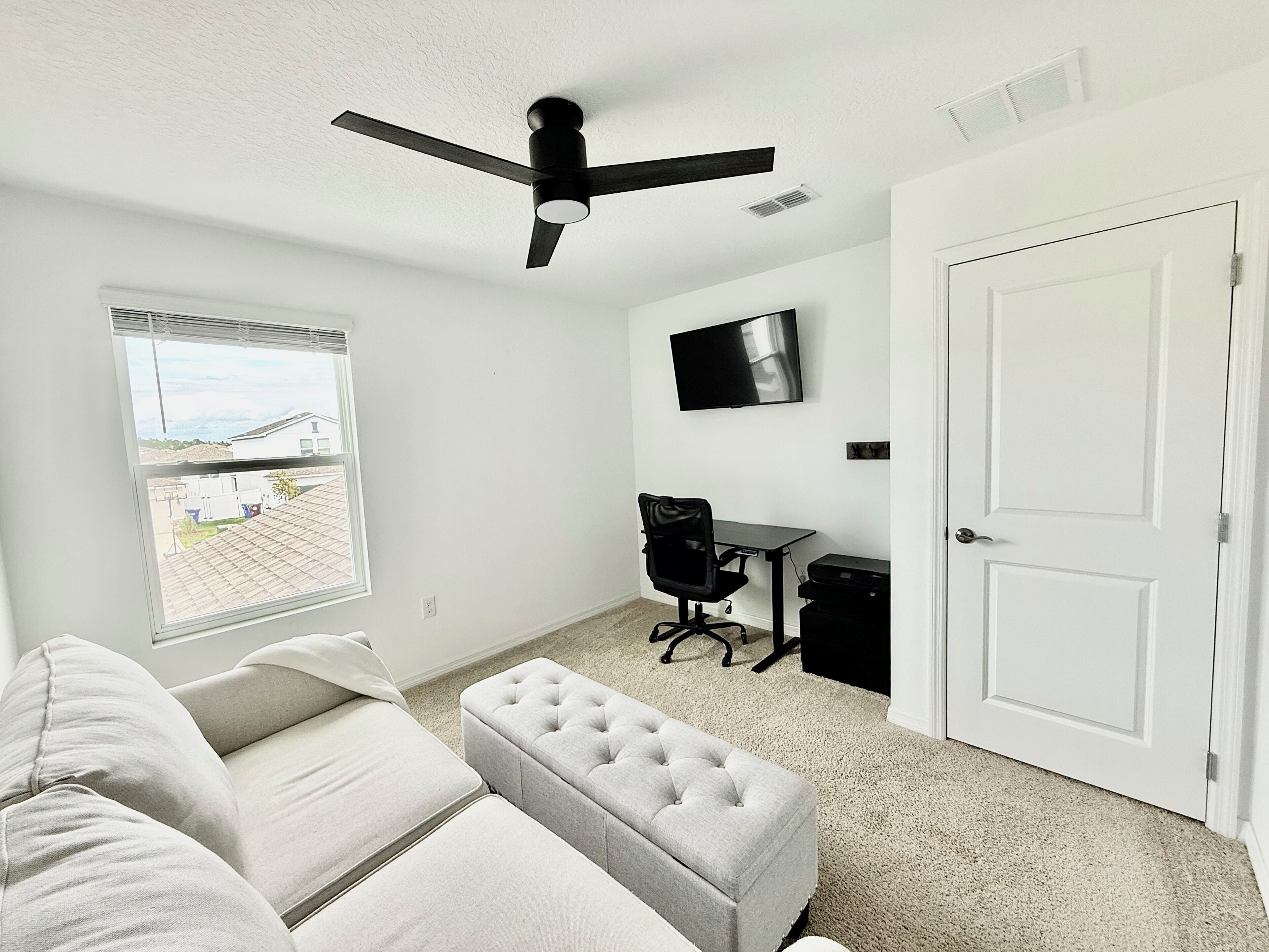 Une pièce dotée d’un ventilateur de plafond, d’un téléviseur fixé au mur, d’un bureau avec une chaise, d’une porte et d’une fenêtre donnant sur un immeuble.