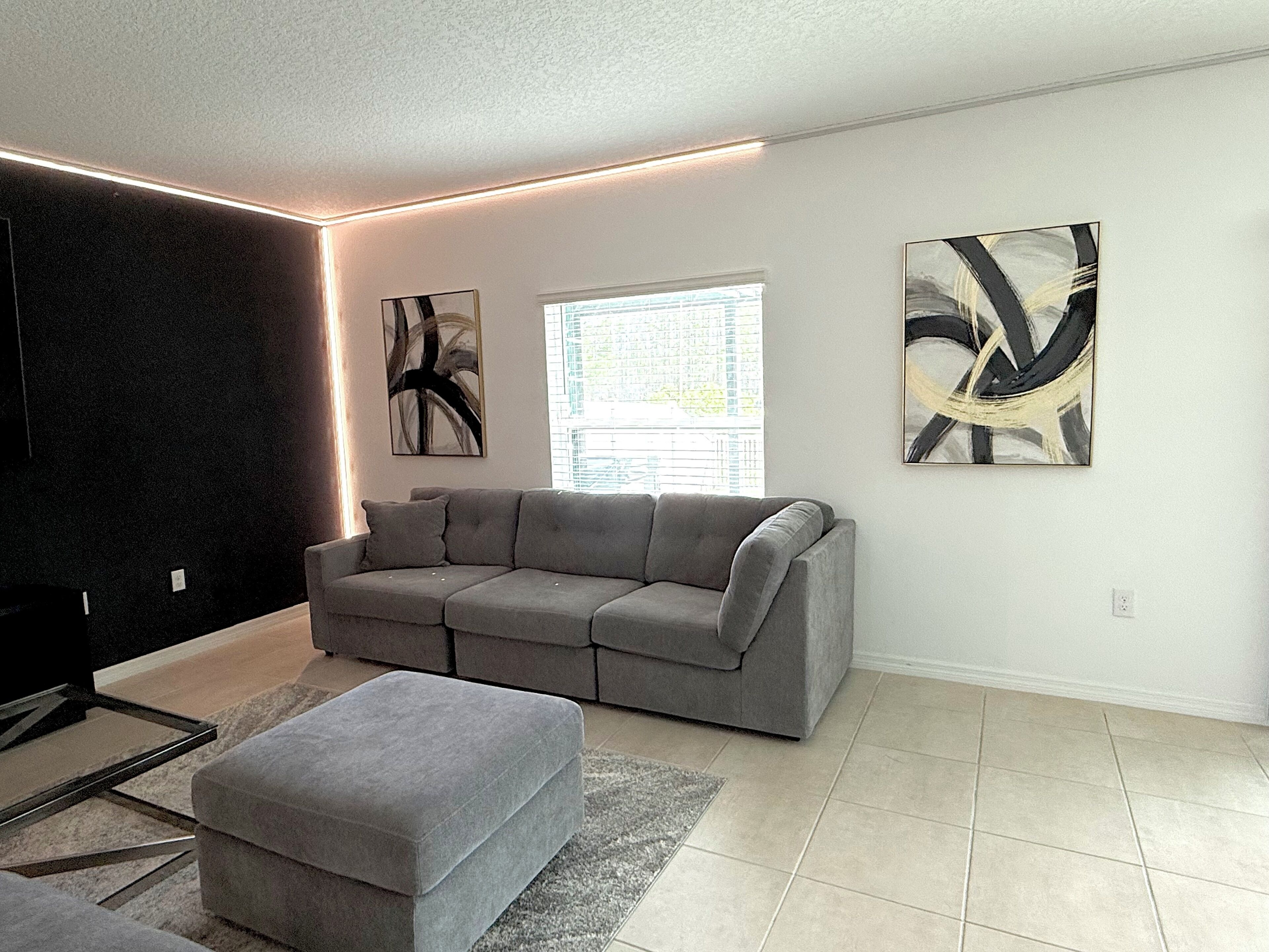 Een moderne woonkamer met een grijze bank, een grijze poef, abstracte wandkunst en een raam met zonwering.