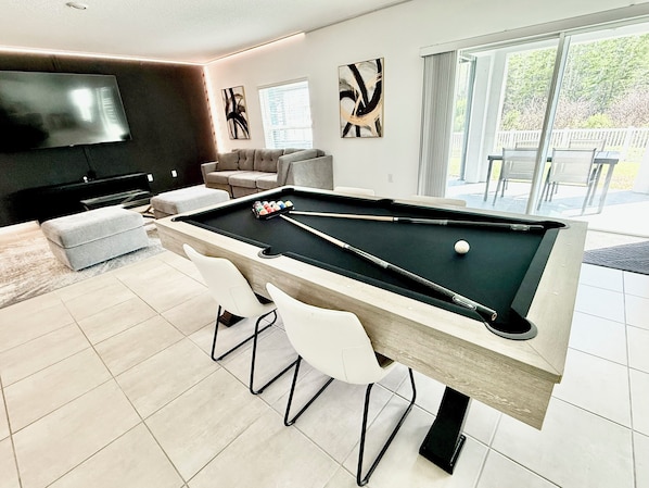 A pool table with a billiard cue, a sofa, a television, and a window with a view of outdoor furniture.