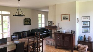 A dining area with a wooden table and chairs, a chandelier, a fireplace, and a living room with a sofa and a television. - Gîte le Moulin Fontaine (Saint-Martin-le-Gaillard)