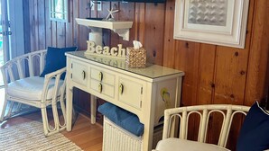 A room with a wooden wall, a flat-screen TV, a white dresser with drawers, wicker chairs, and a woven rug.