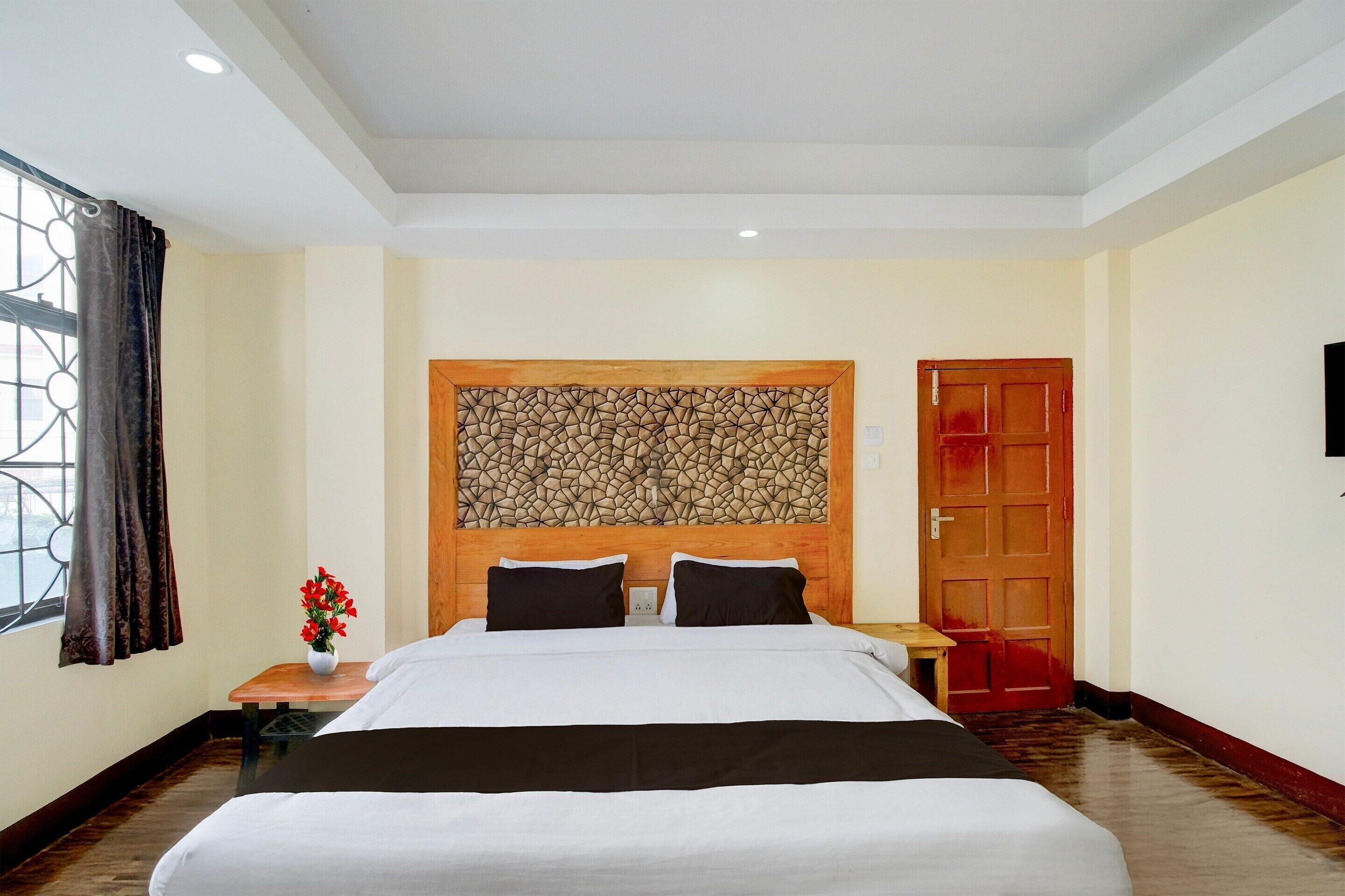 A hotel room with a large bed, a wooden headboard, a small table with a vase of flowers, and a red door.