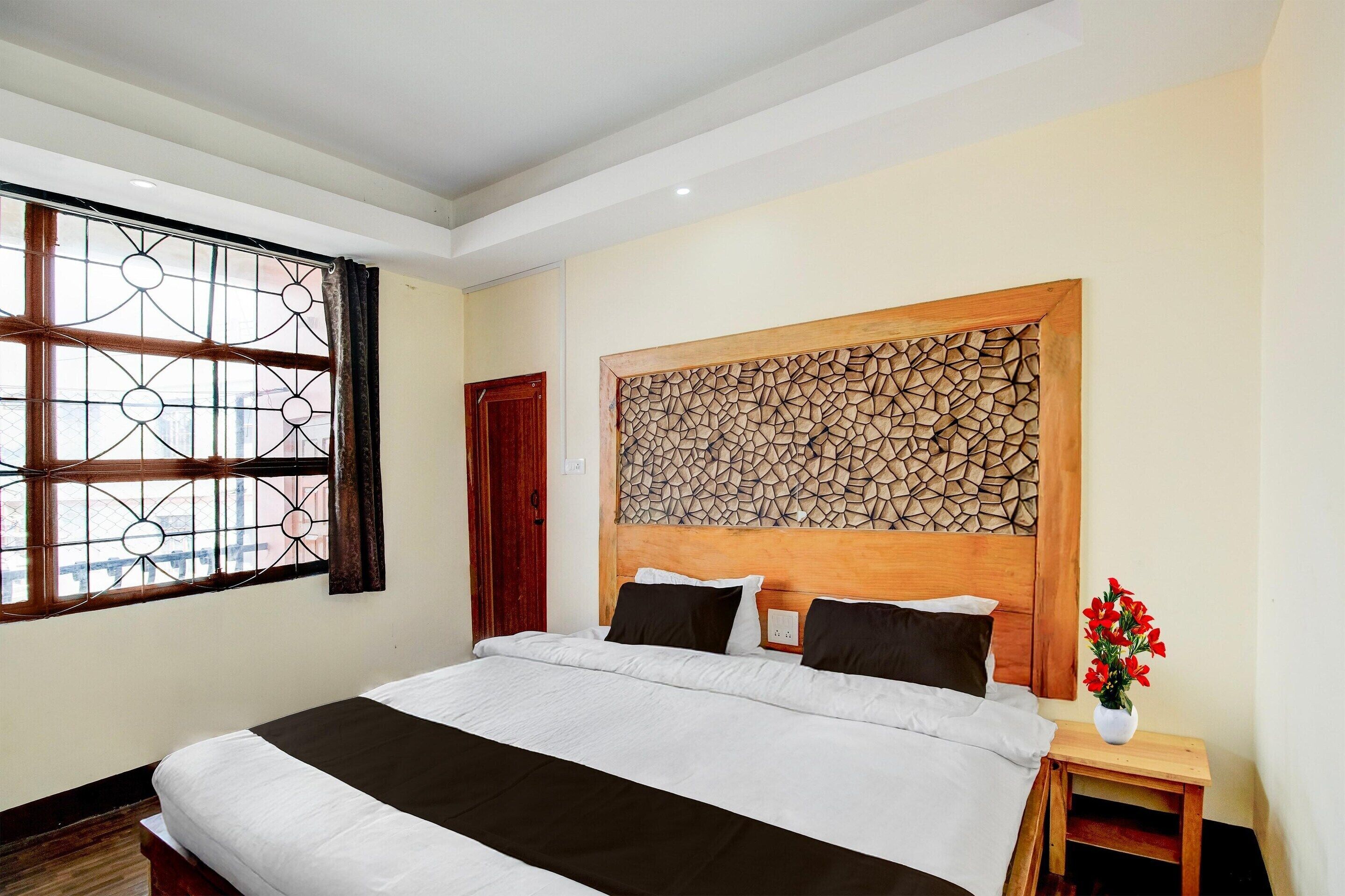 Quarto de hotel com uma cama grande, cabeceira de madeira, janela com cortinas e uma pequena mesa de cabeceira com um vaso de flores.