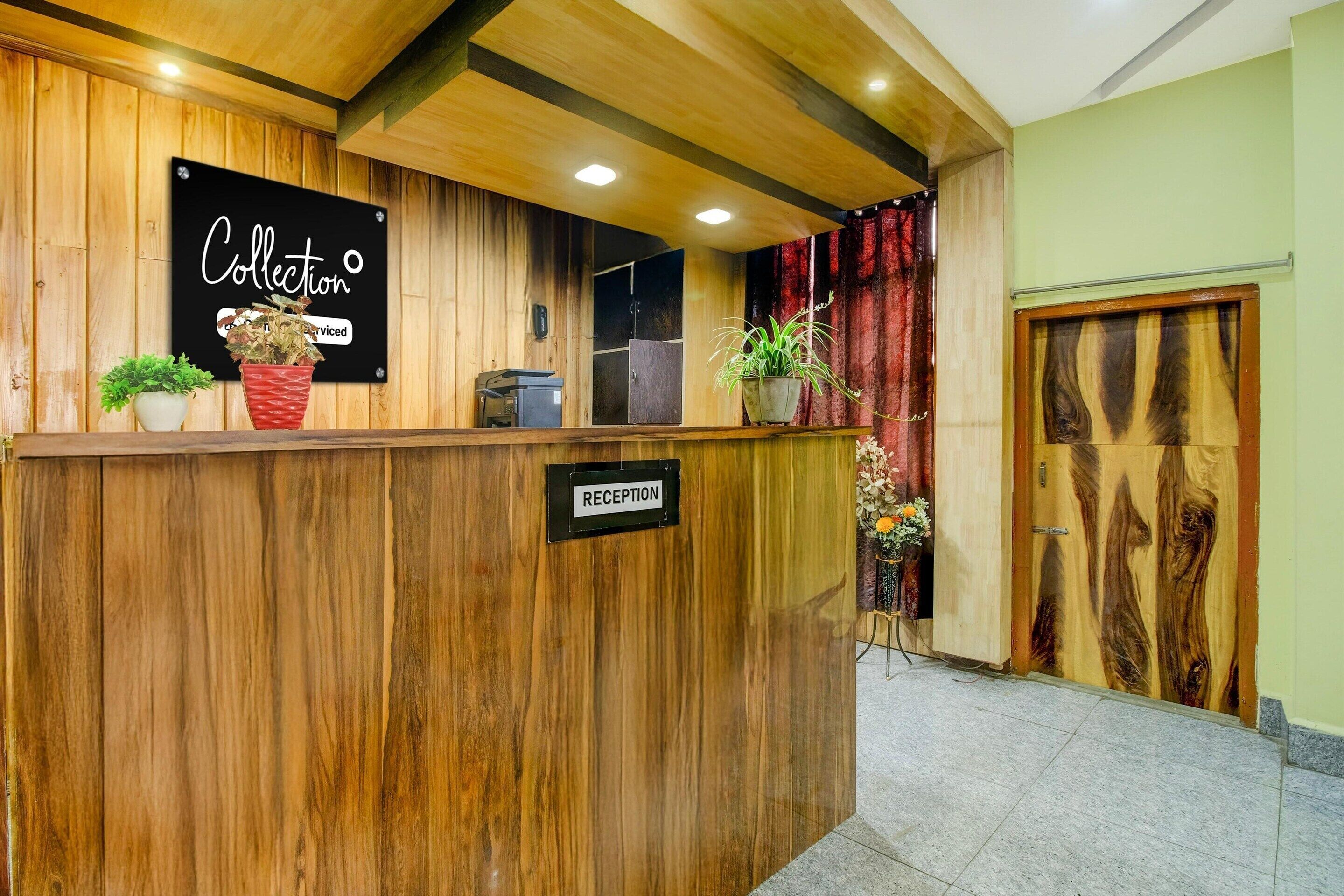 Un mostrador de recepción con un letrero que dice 'Collection' y un letrero de 'RECEPTION' debajo. Hay plantas en macetas sobre el mostrador, y se ve una puerta con diseño de paneles de madera al fondo.