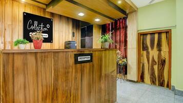Un mostrador de recepción con un letrero que dice 'Collection' y un letrero de 'RECEPTION' debajo. Hay plantas en macetas sobre el mostrador, y se ve una puerta con diseño de paneles de madera al fondo.