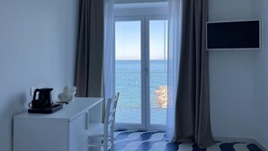 Una habitación con escritorio blanco, cafetera, silla, televisor y vista al mar por un ventanal.