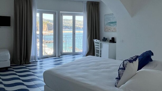 Habitación de hotel con cama, escritorio, silla, televisor y vista al mar a través de amplias ventanas.