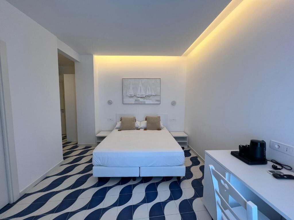A hotel room with a bed, a desk, a chair, a small table, a wall-mounted shelf, and a door leading to another room.