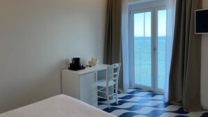 Habitación de hotel con cama, escritorio, silla, cafetera y vista al mar a través de un ventanal grande.