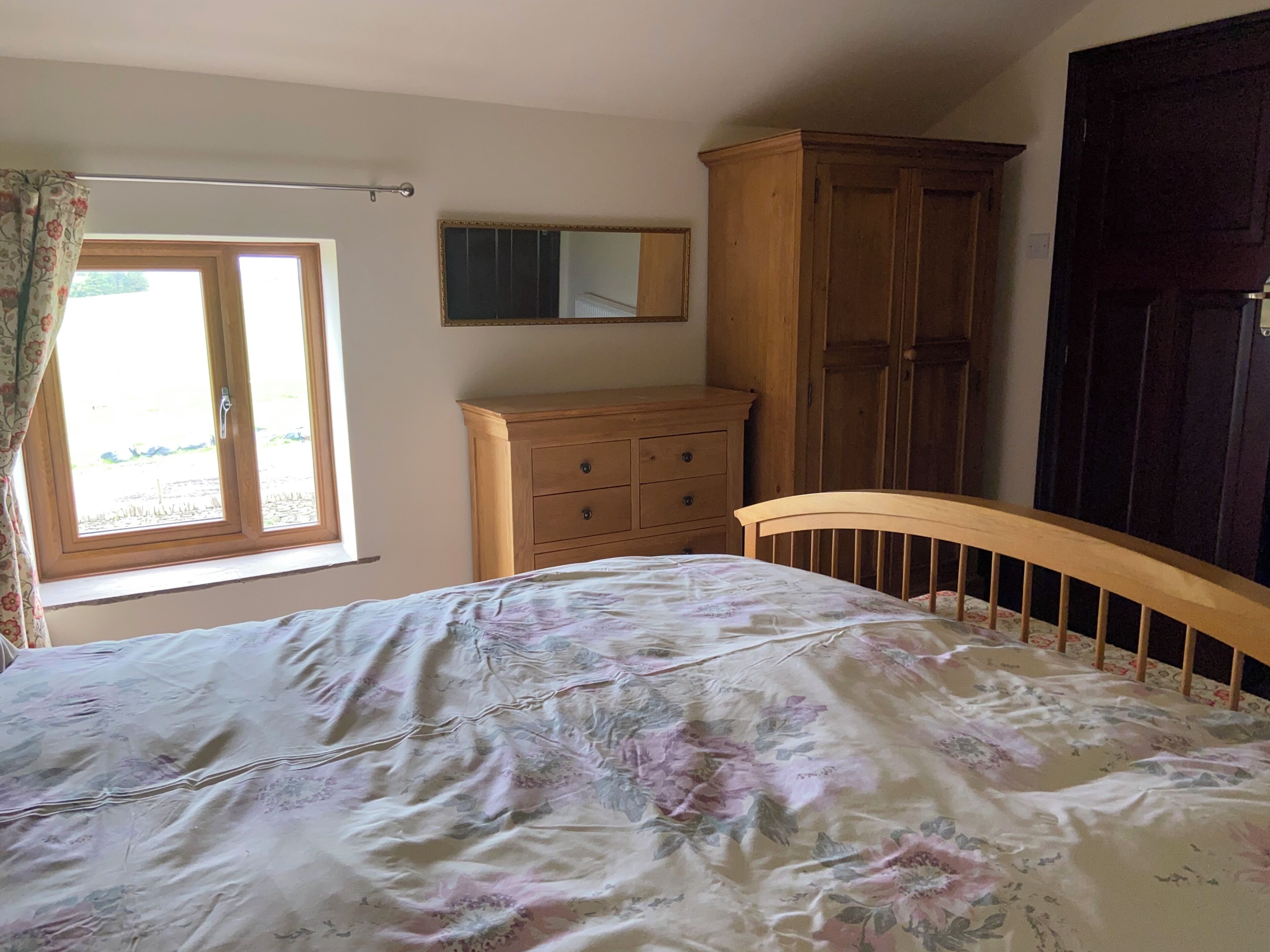 Un dormitorio con cama, cómoda de madera, armario, ventana con cortinas y un espejo en la pared.