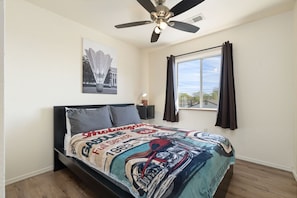Une chambre à coucher avec un lit, un ventilateur de plafond, une fenêtre avec des rideaux et un tableau accroché au mur.