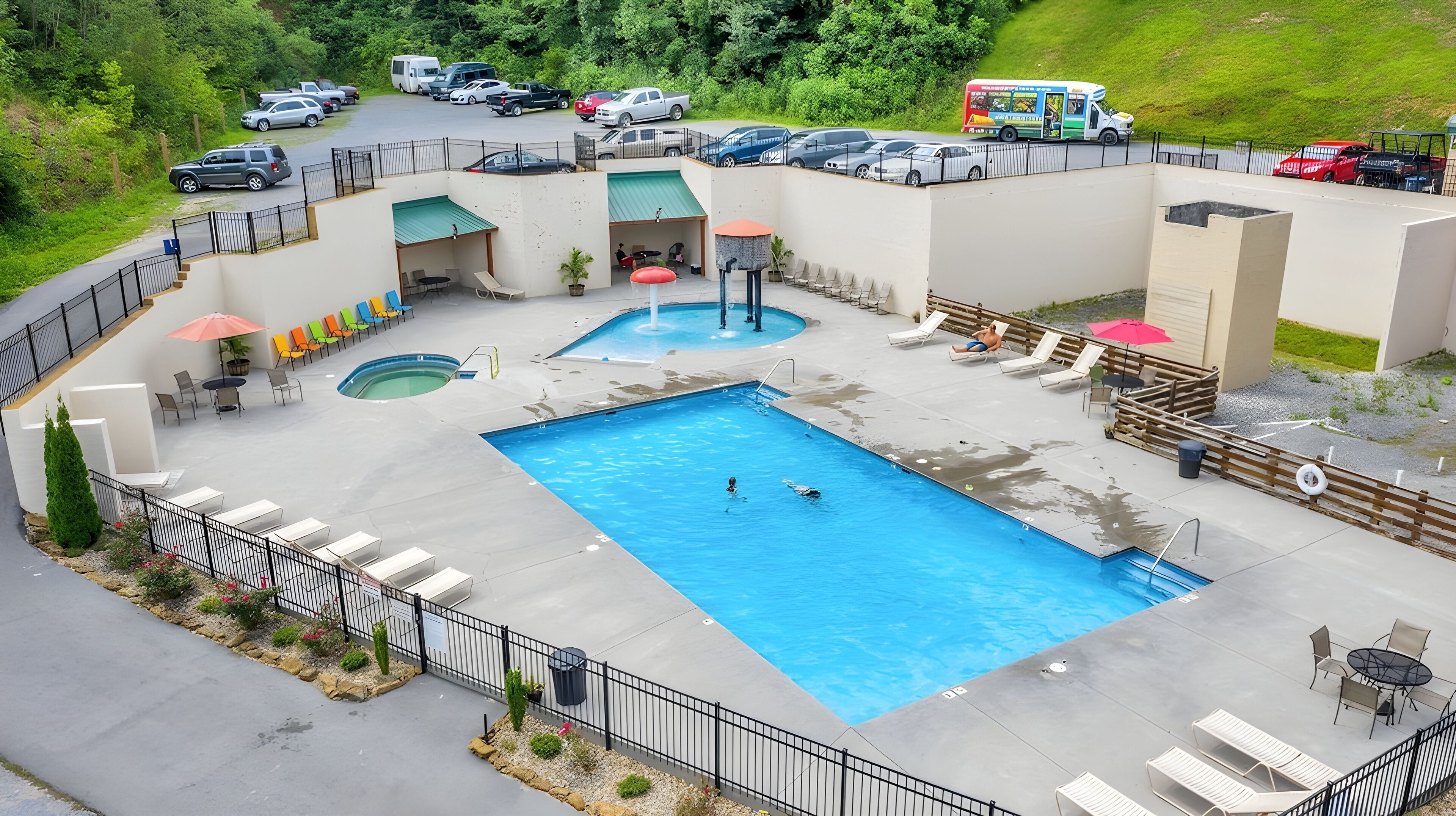 Piscina com espreguiçadeiras e banheira de hidromassagem, cercada por uma área vedada, com um estacionamento visível ao fundo.