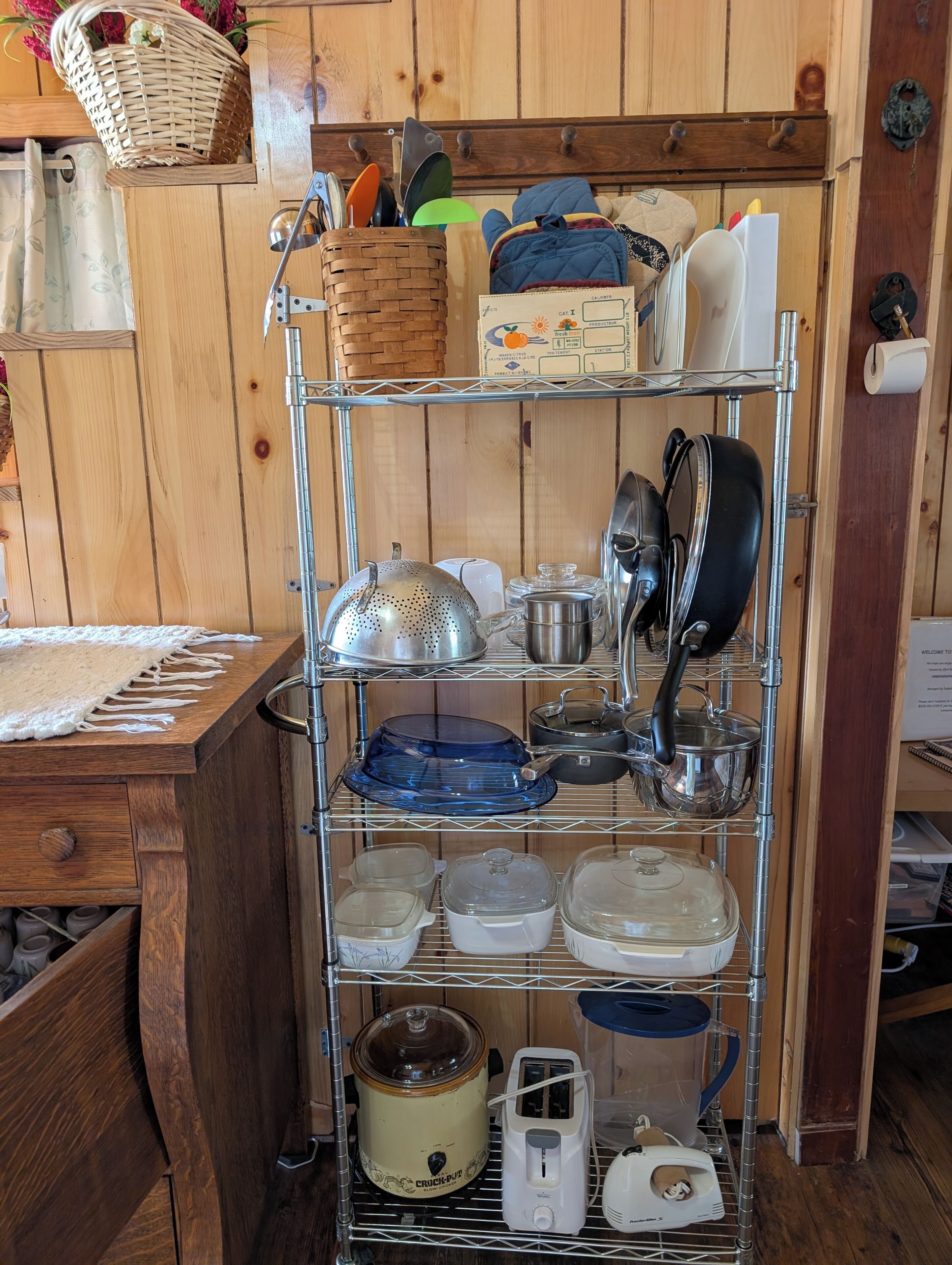 Une cuisine avec un étagère en métal où sont rangés divers pots et casseroles, un autocuiseur, un grille-pain et un panier fixé au mur.