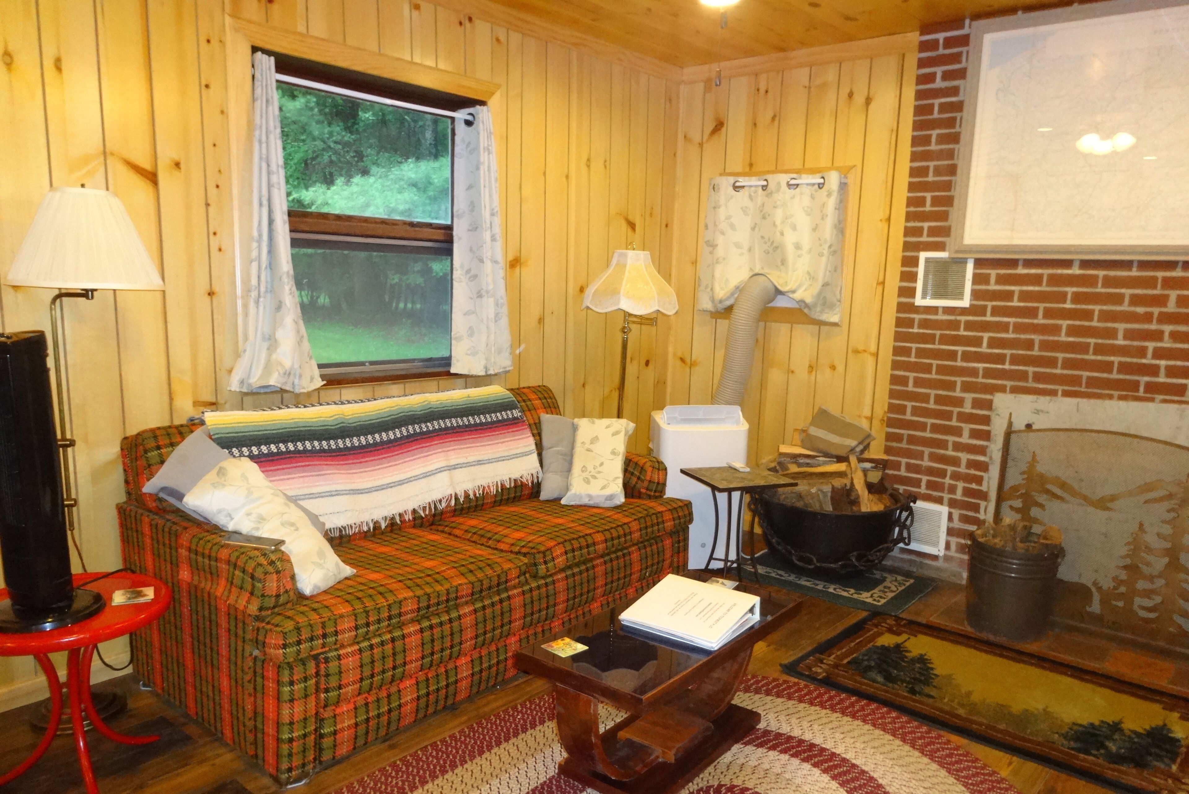 Ein gemütliches Blockhaushaus-Interieur mit einem karierten Sofa, einem roten Sessel, einem Holztisch, einem Kamin und einem Fenster mit Vorhängen.