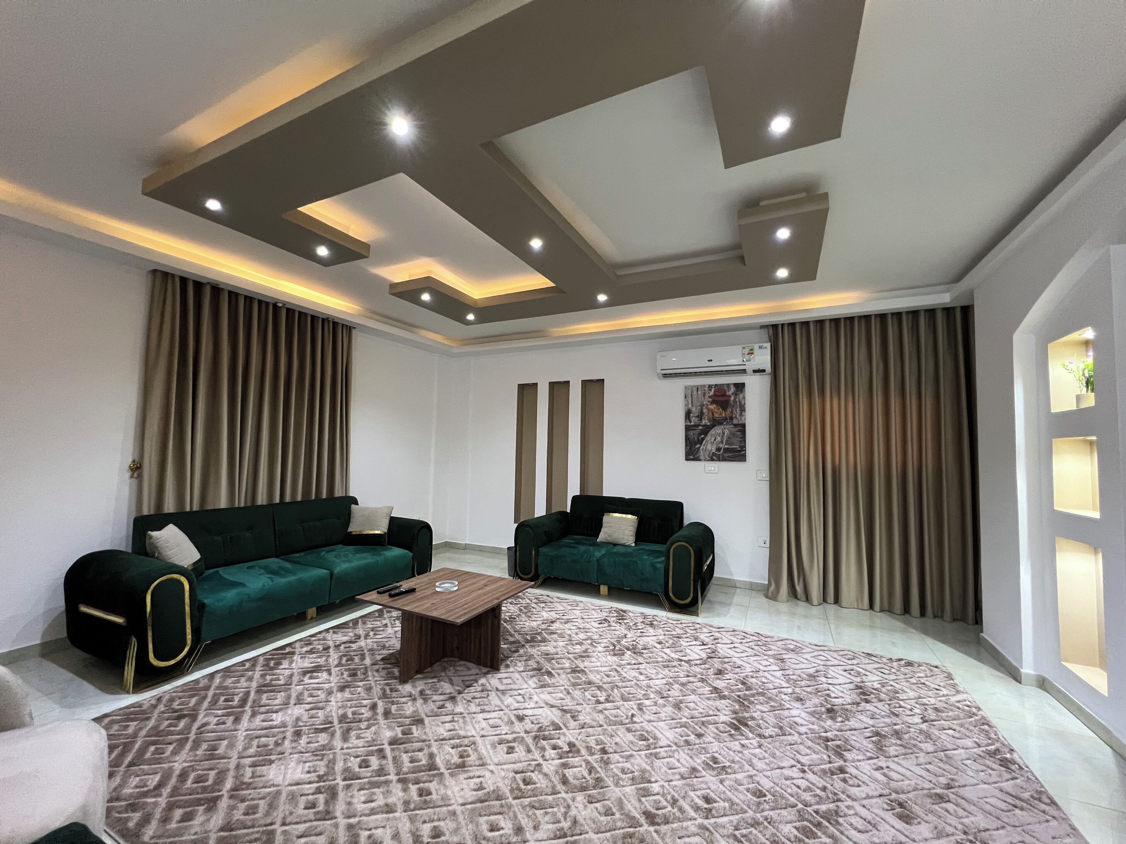 Un salon moderne comprenant un ensemble de canapés, une table basse, un tapis à motifs, un éclairage encastré au plafond et un appareil de climatisation.