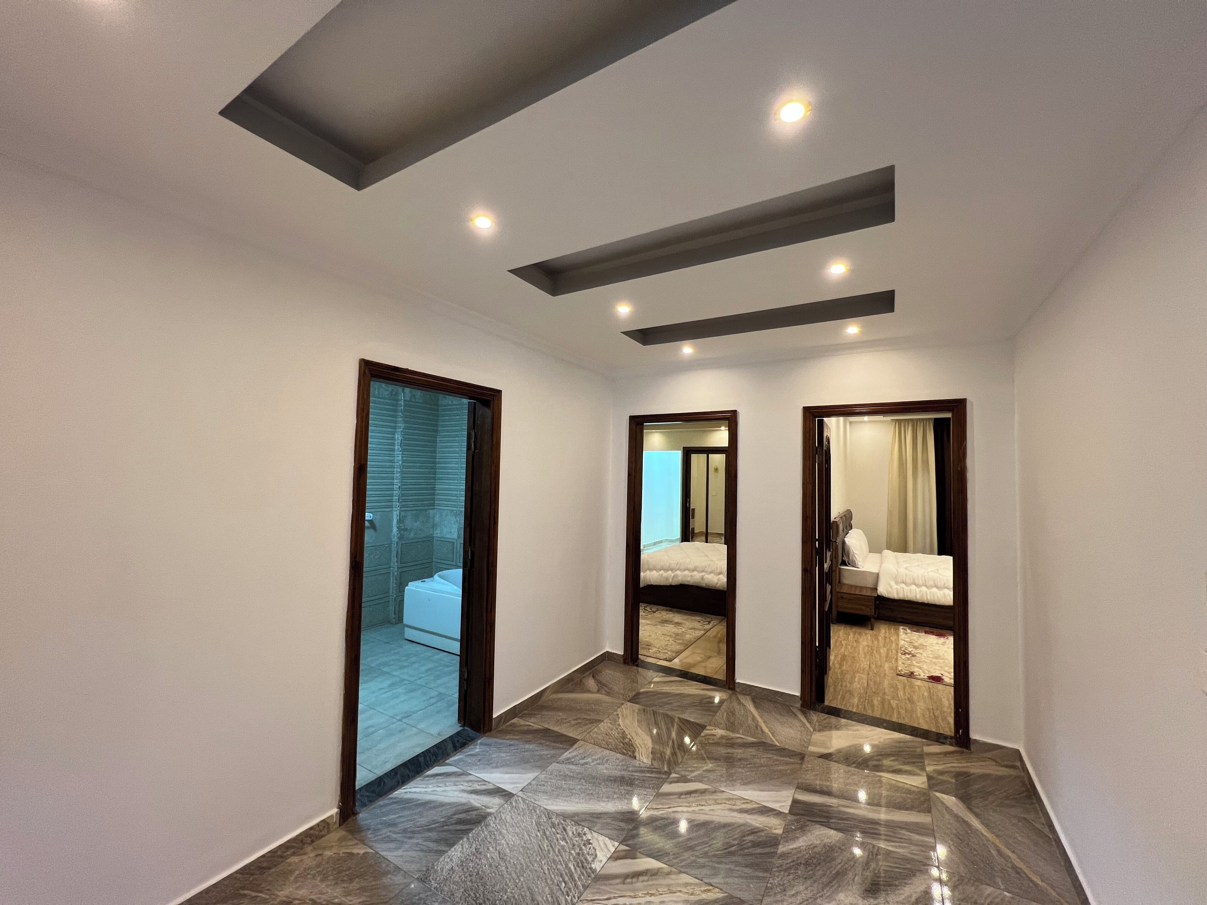 A hallway with a tiled floor, a door leading to a bathroom, and a mirror reflecting a bedroom.