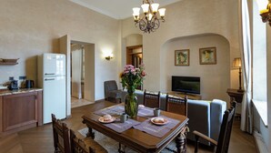 Un coin repas avec une table en bois, des chaises, un lustre, un téléviseur et, au fond, une cuisine.