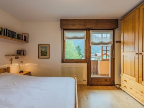 Ein Schlafzimmer mit einem Bett, einem Holzschrank, einem Fenster mit Blick auf ein Haus, einer Heizung und einem Bücherregal mit Büchern.