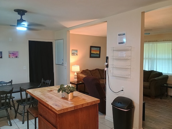 A compact living space with a wooden dining table, chairs, a small kitchen area, a sofa, and a trash bin.