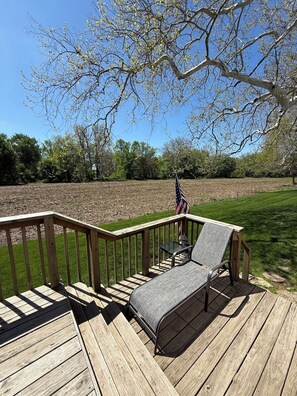 A wooden deck with a metal table and two chairs, an American flag, and a view of a field and trees. - Welcome to your quaint peaceful getaway in the heart of small-town Indiana! (Roann)