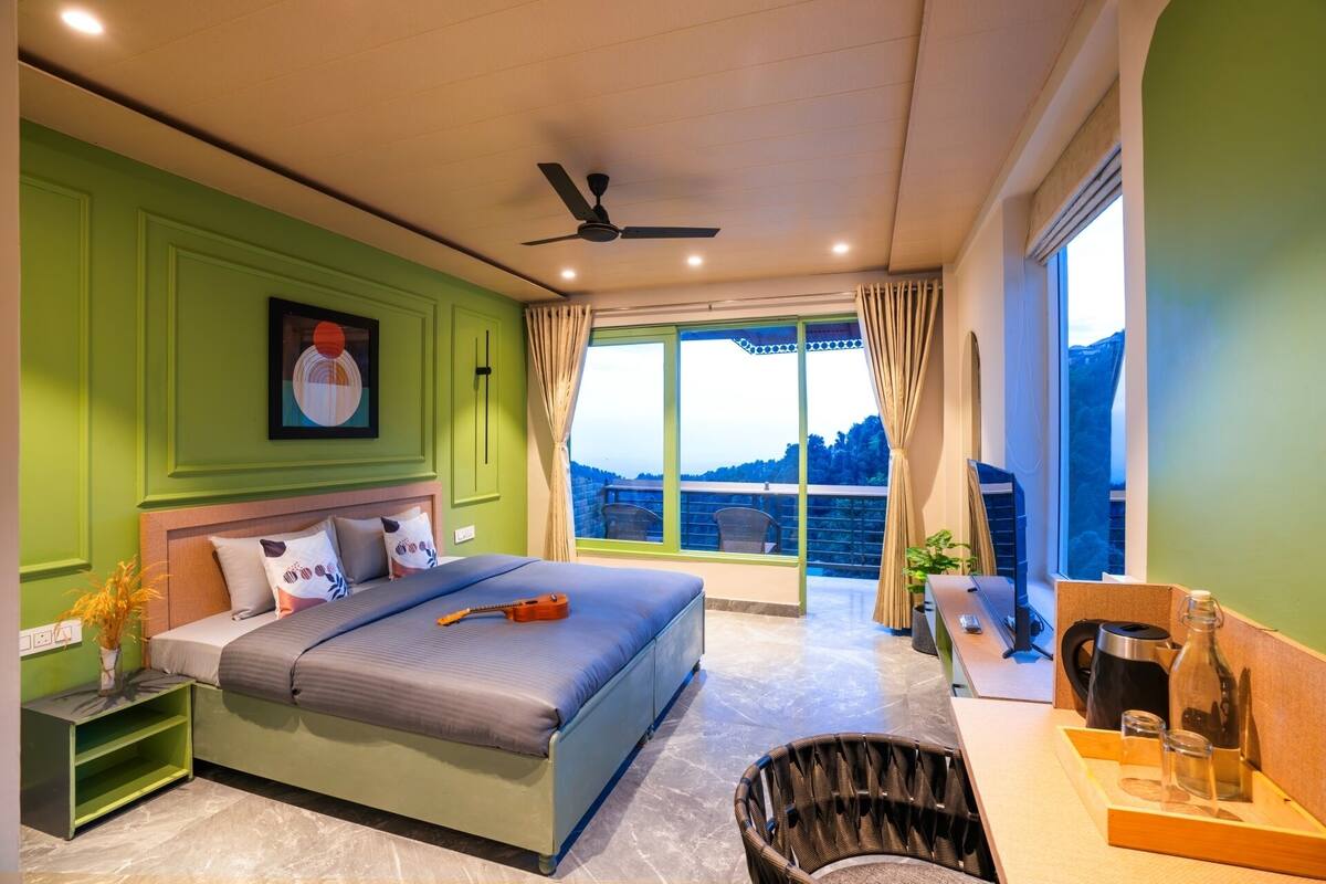 A bedroom with a bed, a ceiling fan, a window with curtains, a view of the outdoors, and a small table with a kettle and glasses.